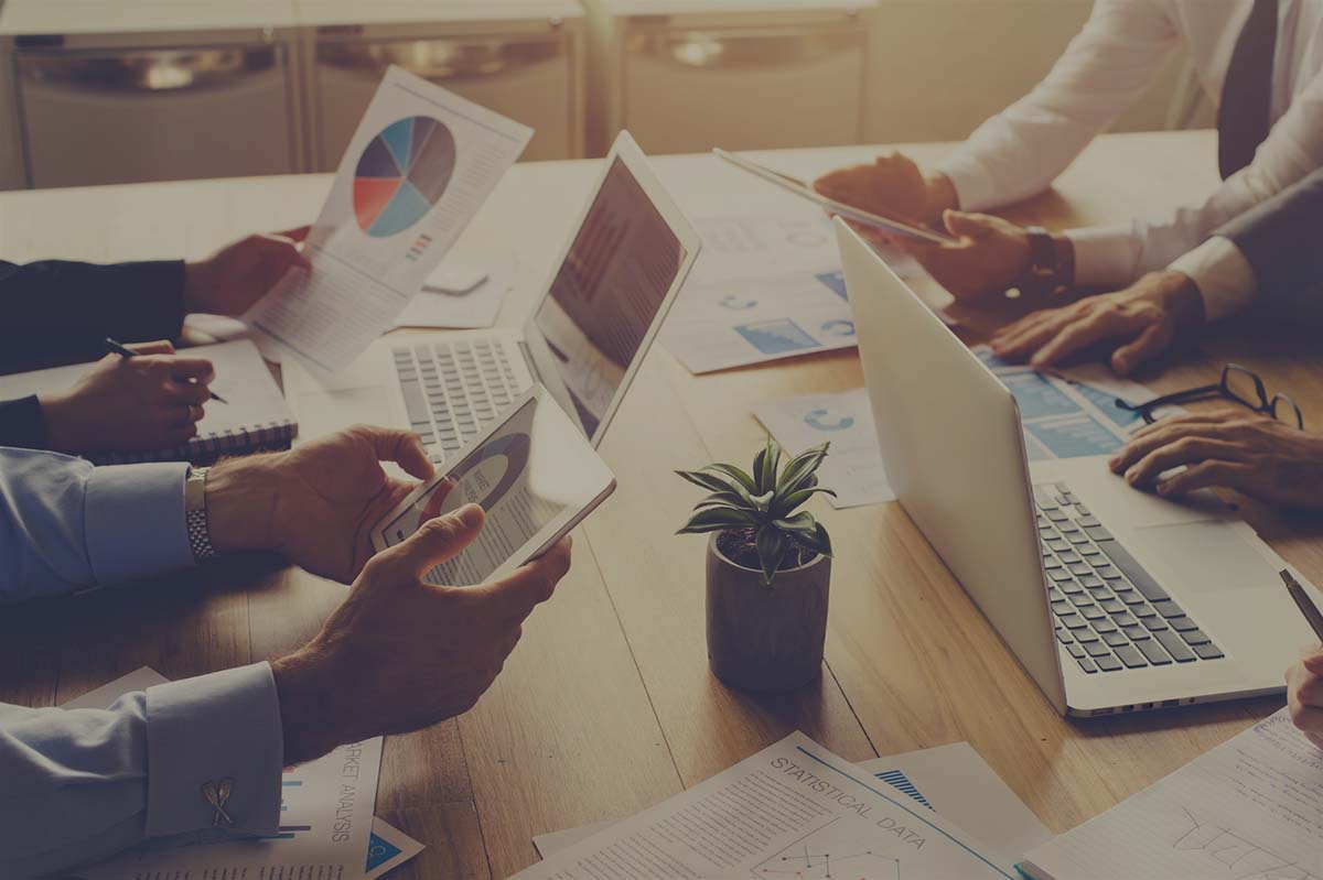 Group of people meeting with technology. They are around a conference table with digital tablets and laptops. There are marketing and strategy documents on the table